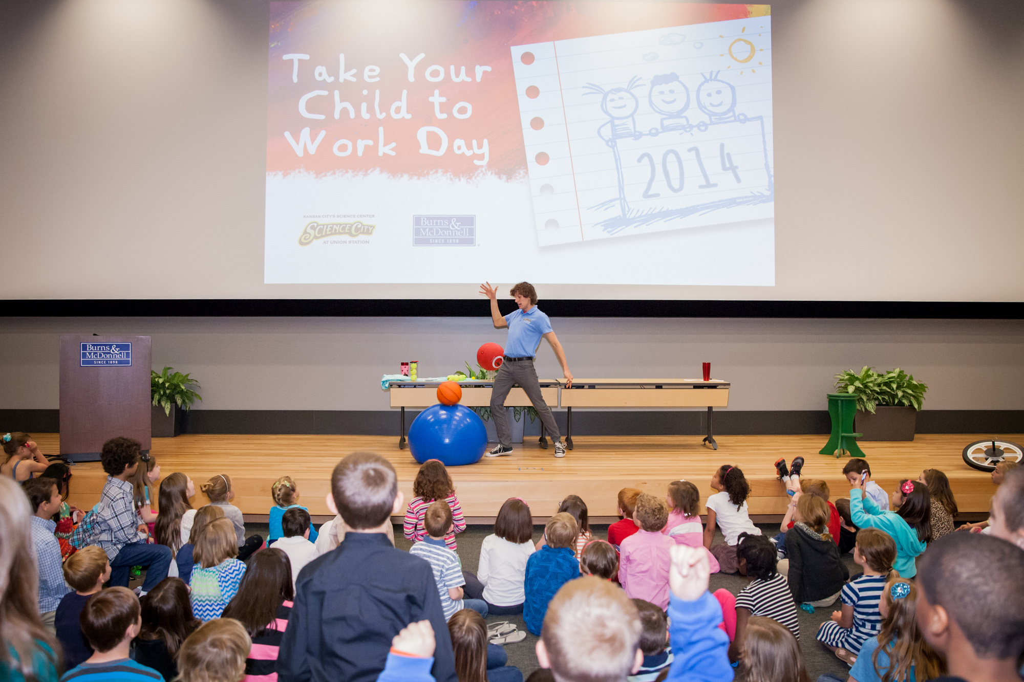 An image showing a man presenting to a room full of children. The man is showing a science experiment where one ball is dropped on top of another ball to see how high it will bounce.