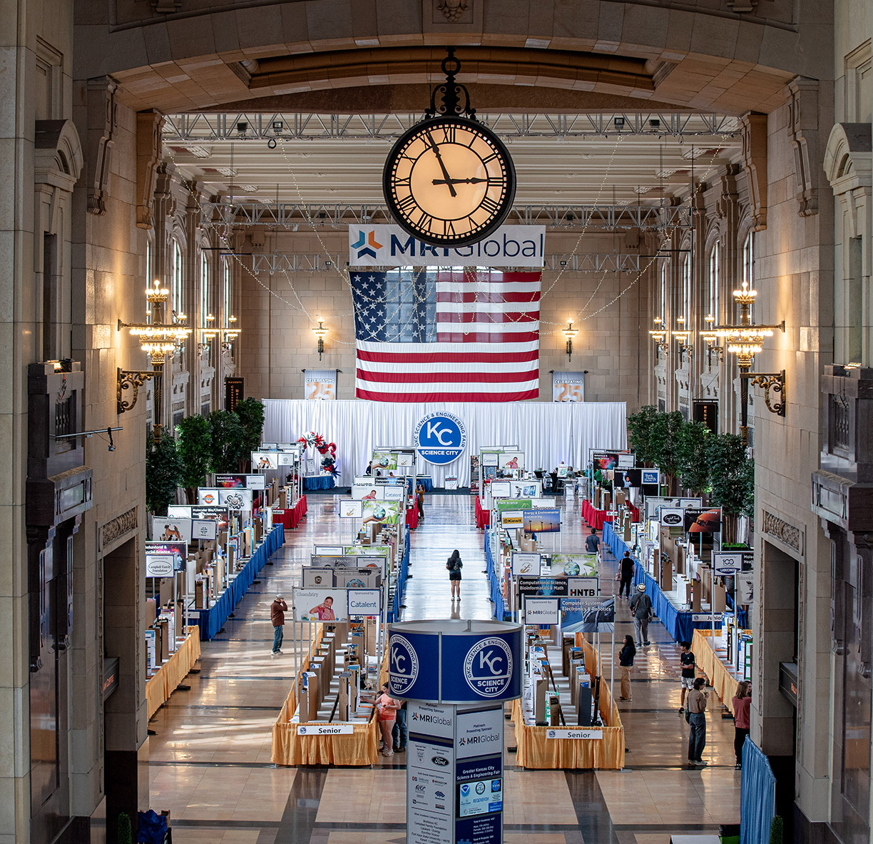 A photo from the 2025 Greater Kansas City Science and Engineering Fair. Showing a stage with a backdrop behind it, guest seated in chairs, and the MRI Global Logo.
