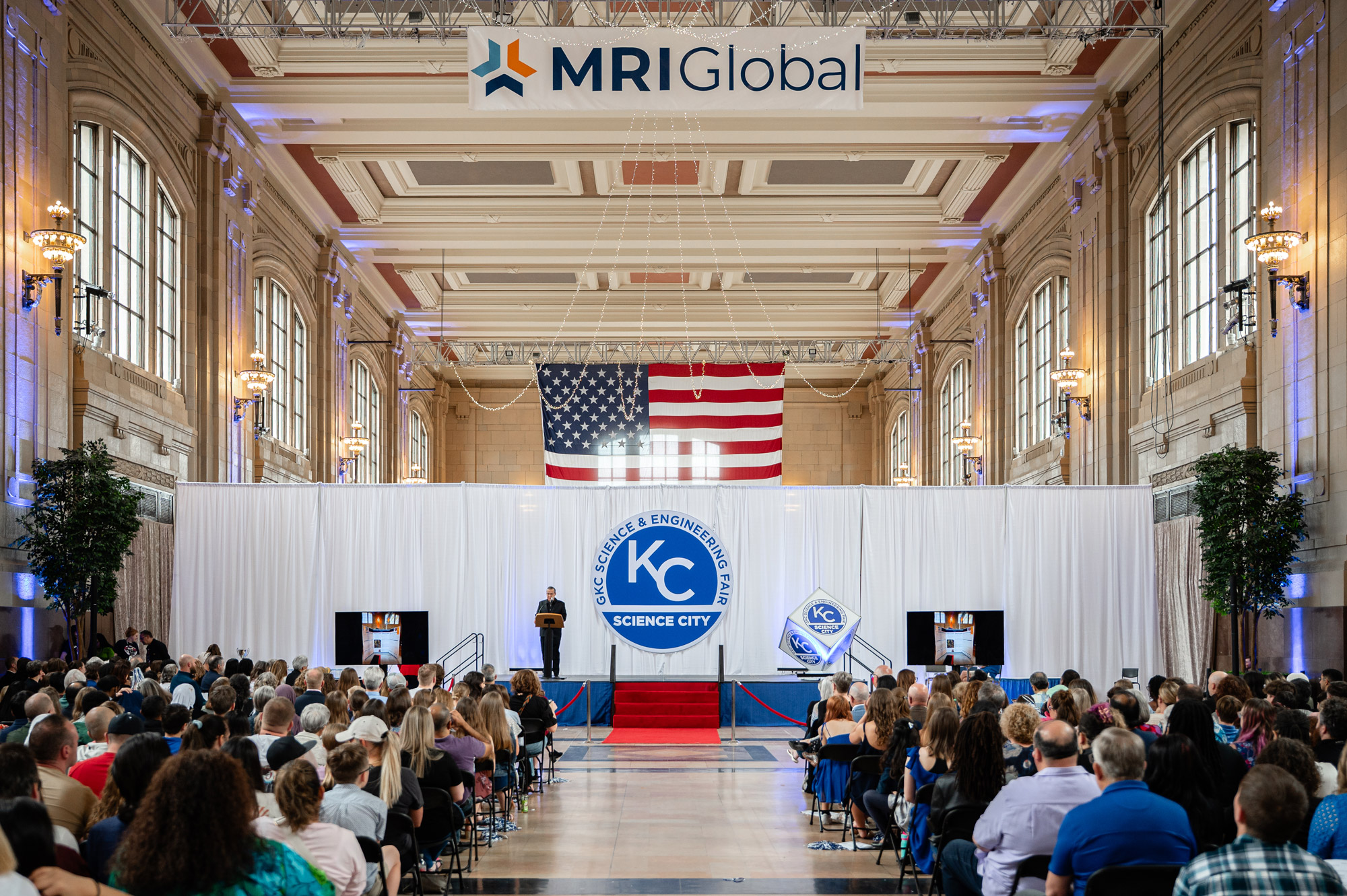 A photo from the 2025 Greater Kansas City Science and Engineering Fair. Showing a stage with a backdrop behind it, guest seated in chairs, and the MRI Global Logo.